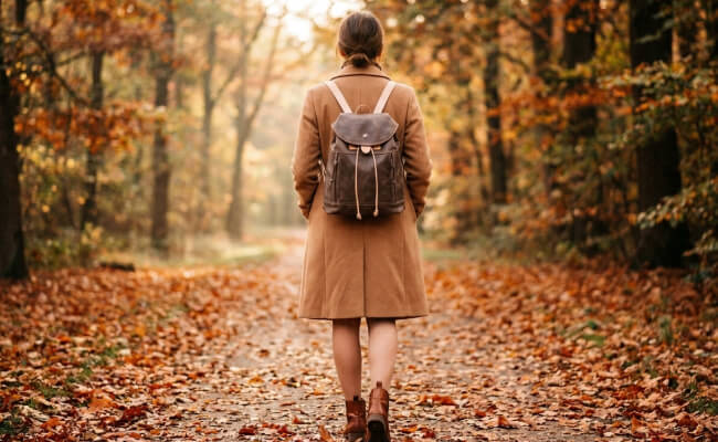 A woman wearing a brown and beige leather backpack on her back, walking through an autumn park.