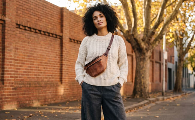 A woman wearing a cognac brown leather fanny pack worn crossbody over a chunky knit sweater and wide-leg trousers.