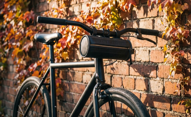 A black leather cylindrical bicycle bag strapped to the handlebar of a matte black city bicycle.