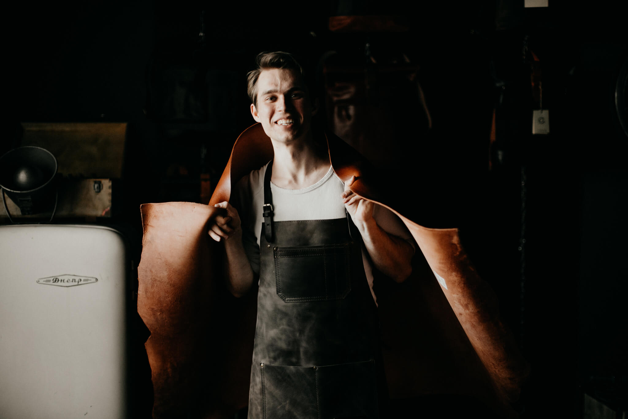 Smiling leather artisan holding a large leather hide while wearing a workshop apron.