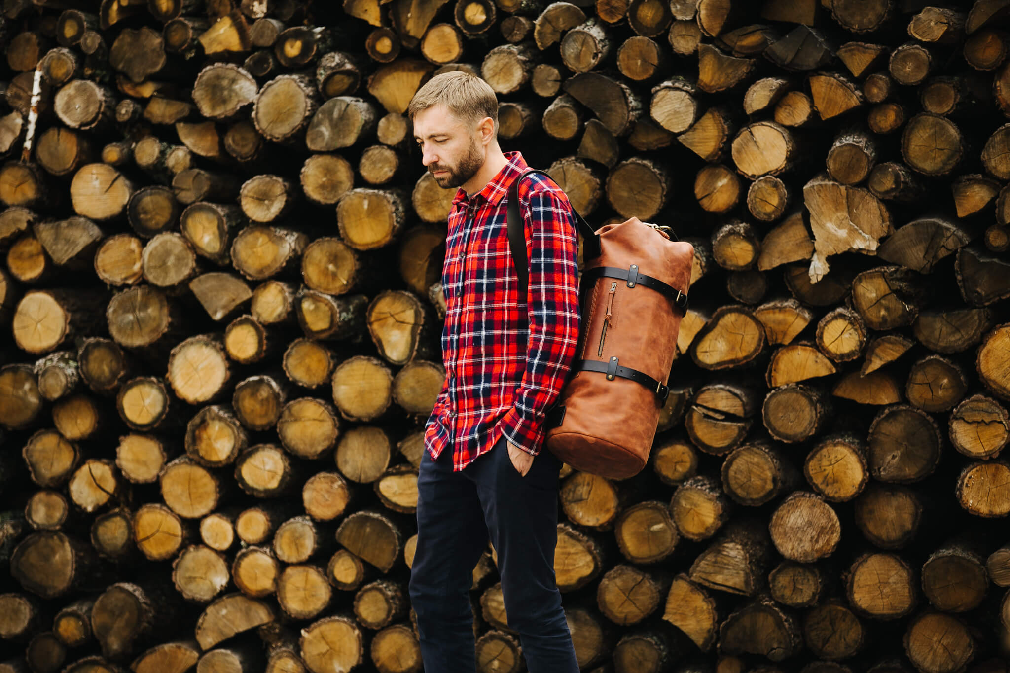 Man wearing Kruk Garage DuffelPack SE010 leather bag against stacked firewood backdrop, showcasing rugged summer travel style.