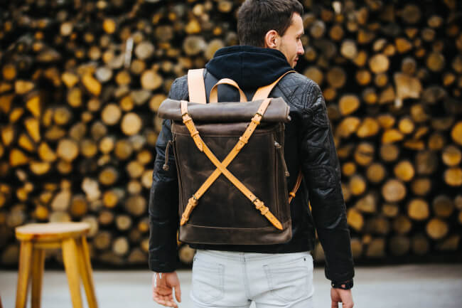 Man wearing Kruk Garage leather Crossroad backpack, walking with a rustic woodpile background, showcasing stylish travel accessories.
